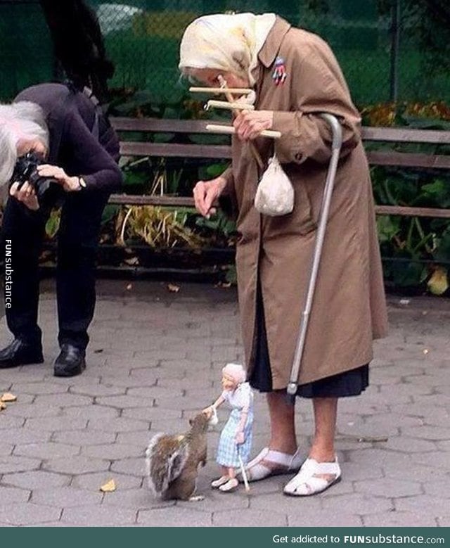 Old woman uses a marionette of herself to feed squirrels in the park